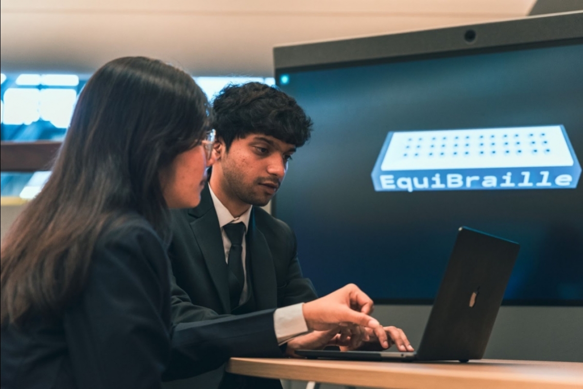 Two students work on a laptop while in the background a 3D render of a board with "EquiBraille" written on it is displayed