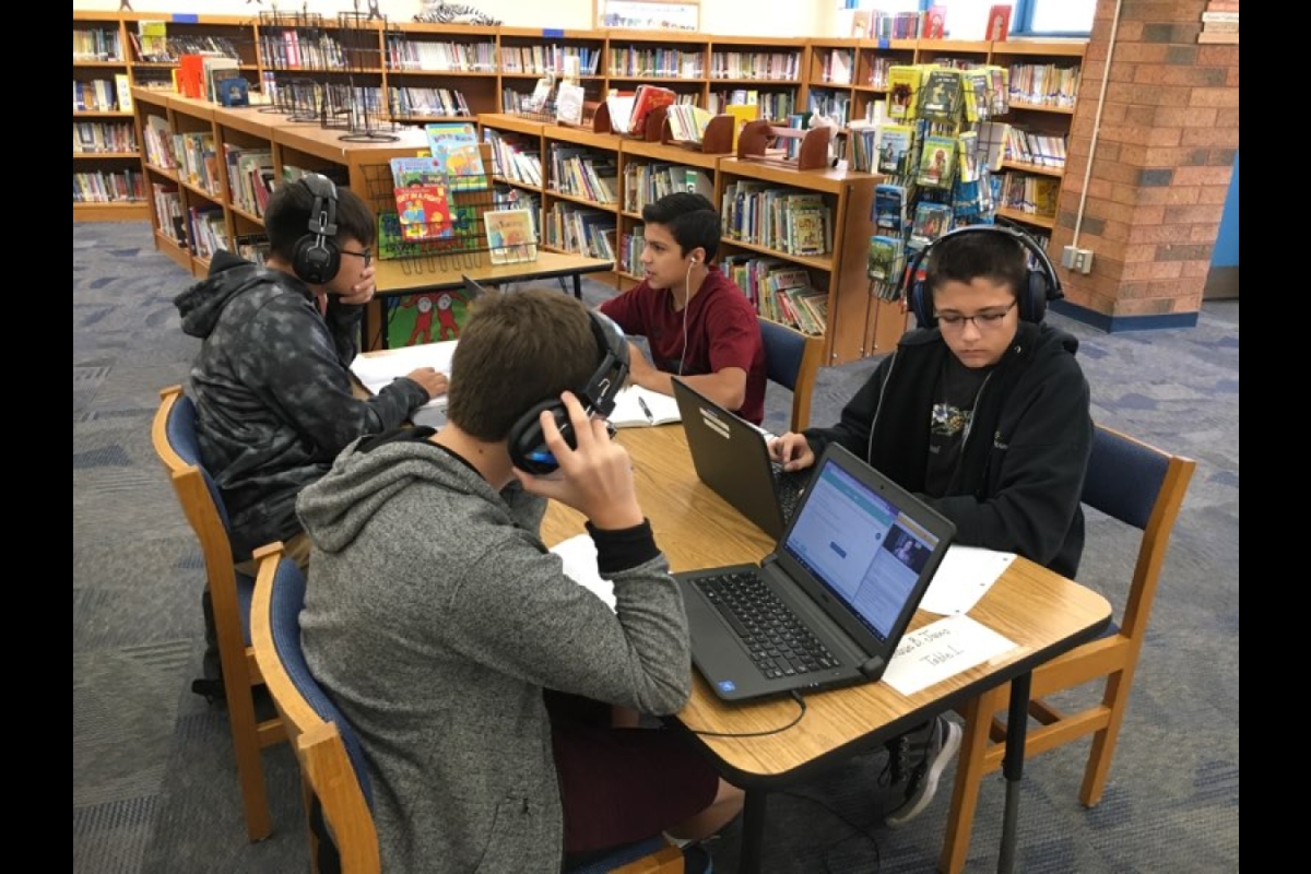 Four middle school students sit at a table in a school library logged in to a digital class, wearing headphones in front of laptops