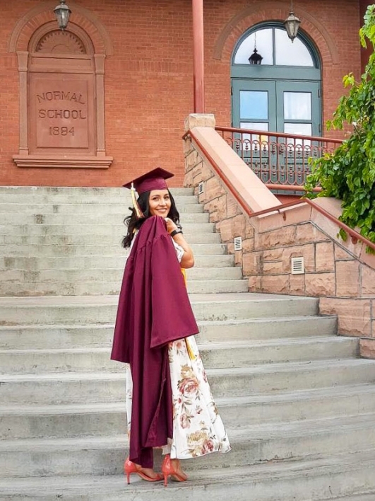 ASU's College of Health Solutions graduate Dulce Vivas Cruz wears her cap and gown as she stands on the staircase at Old Main on ASU's Tempe campus and looks back over her shoulder and smiles