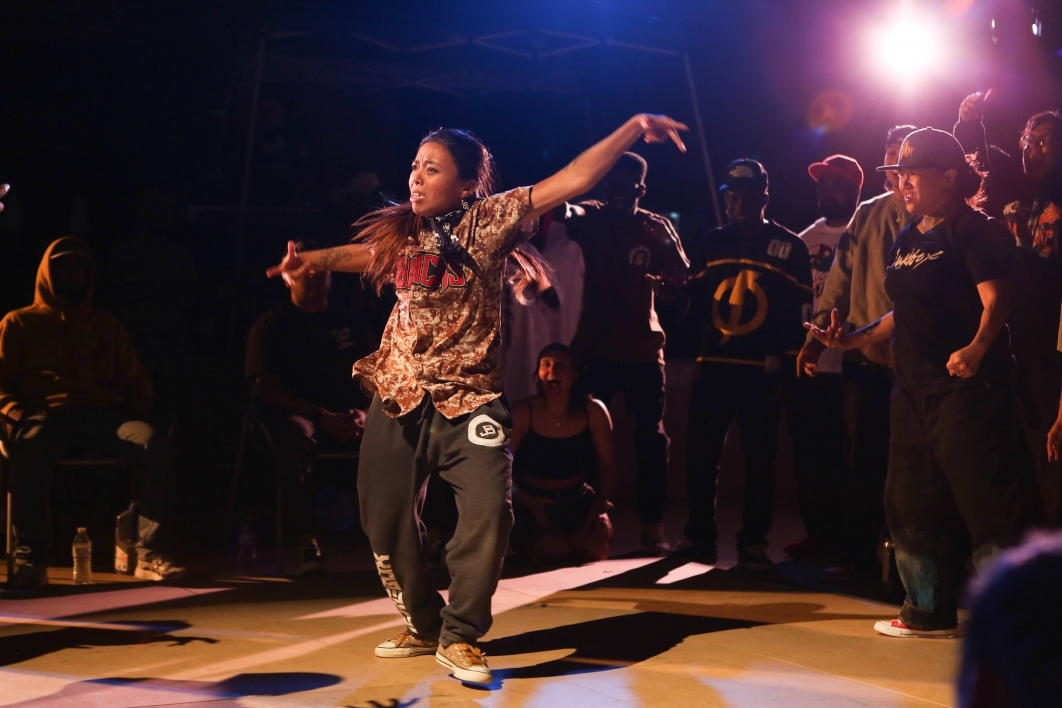A person in the middle of a circular crowd performs a hip-hop dance.