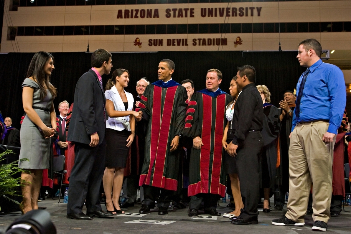 Former U.S. President Barack Obama stands on stage with ASU President Michael Crow wearing graduation regalia and shaking hands with students.