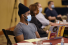A young Black man listens to a panel discussion. He is wearing a gray V-neck shirt, black beanie and a blue surgical face mask. He is sitting at a round table with a laptop in front of him covered in maroon and gold ASU stickers.