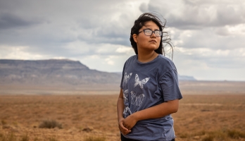 A young Native American woman looks off to the right with a desert landscape stretching behind her of scrub brush leading to mountains in the distance