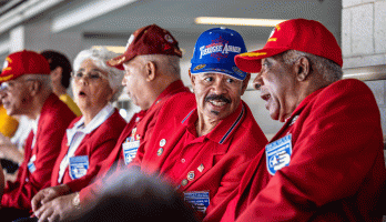 Older men in Tuskegee Airmen hats chat while sitting in stadium seats.