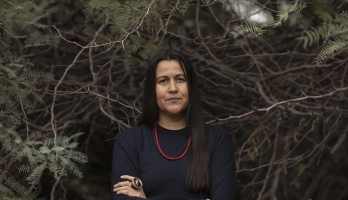 ASU Pulitzer Prize-winner Natalie Diaz wears a black shirt and red and black beaded necklaces as she stands with arms crossed against a backdrop of foliage.