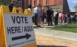 Sign reading "Vote here/aqui" in the foreground and a line of people in the background.