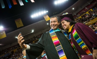 Young student posing with a program director at ASU's Hispanic convocation. Both wear graduation regalia, with the student in a maroon cap and gown and the director in a black gown, and both wear bright, multi-colored stoles.