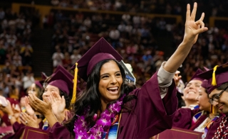 Woman wearing cap and gown at an ASU graduation ceremony. She is smiling widely and making the pitchfork gesture.