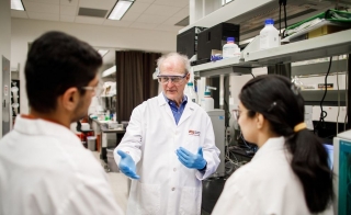 Bruce Rittmann, wearing a white lab coat and goggles, talks to two fellow researchers. 