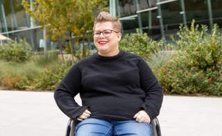 Photo of Rachel Welshans. She sits in a wheel chair and is wearing a plain black long sleeved shirt and jeans. She wears square glasses and red lipstick. 