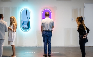 Man standing in front of an art piece featuring two mirrors, one lit by a blue neon light and one lit by a purple neon light.