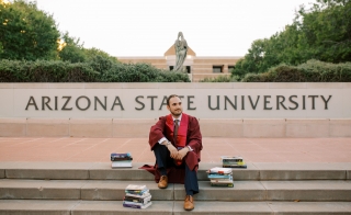Bryson Brown sitting on steps in front of Arizona State University in graduation gown surrounded by books