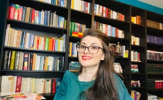 Gina Scarpete Walters smiles at the camera. She is wearing a teal blouse or dress. She has long brown wavy hair that is pulled over one shoulder. She is wearing black framed glasses and red lipstick. Behind her are many black shelves filled with books.