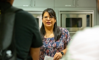 Woman with glasses and long dark hair standing in a demonstration kitchen speaking to students who are seen from behind.