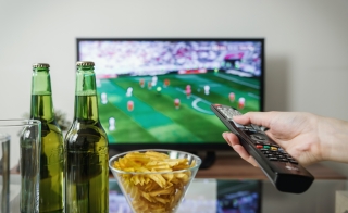 Hand pointing a remote at a screen with football playing on it. There is a coffee table in the foreground with chips and drinks on it.