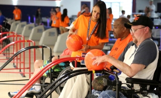 A woman holding a bowling ball speaks to two men in wheelchairs holding bowling balls.