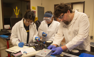 Three students in lab gear examine their car battery using various instruments.
