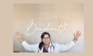 Tanya Carroccio wears a white nurse practitioner coat and stethoscope with her arms raised in the air underneath a sign that says Benehealth in cursive 