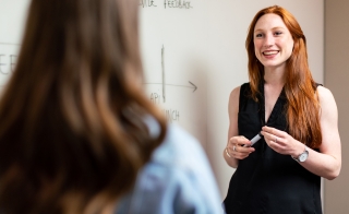 Woman standing next to a whiteboard and smiling while looking at another woman facing her, seen from behind.