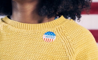 An African-American woman wears a "I Voted" sticker on a yellow sweater