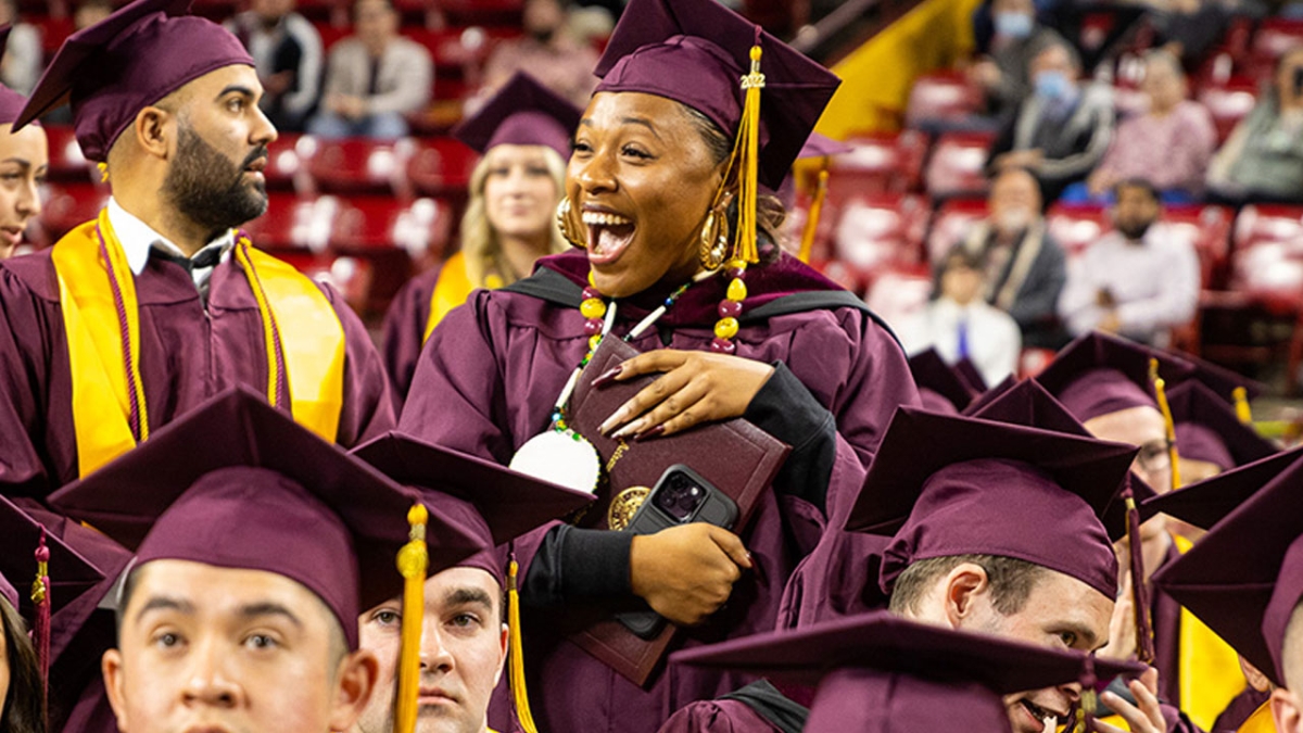 Students wearing maroon and gold caps and gowns, smiling widley at the fall 2022 Cronkite convocation.