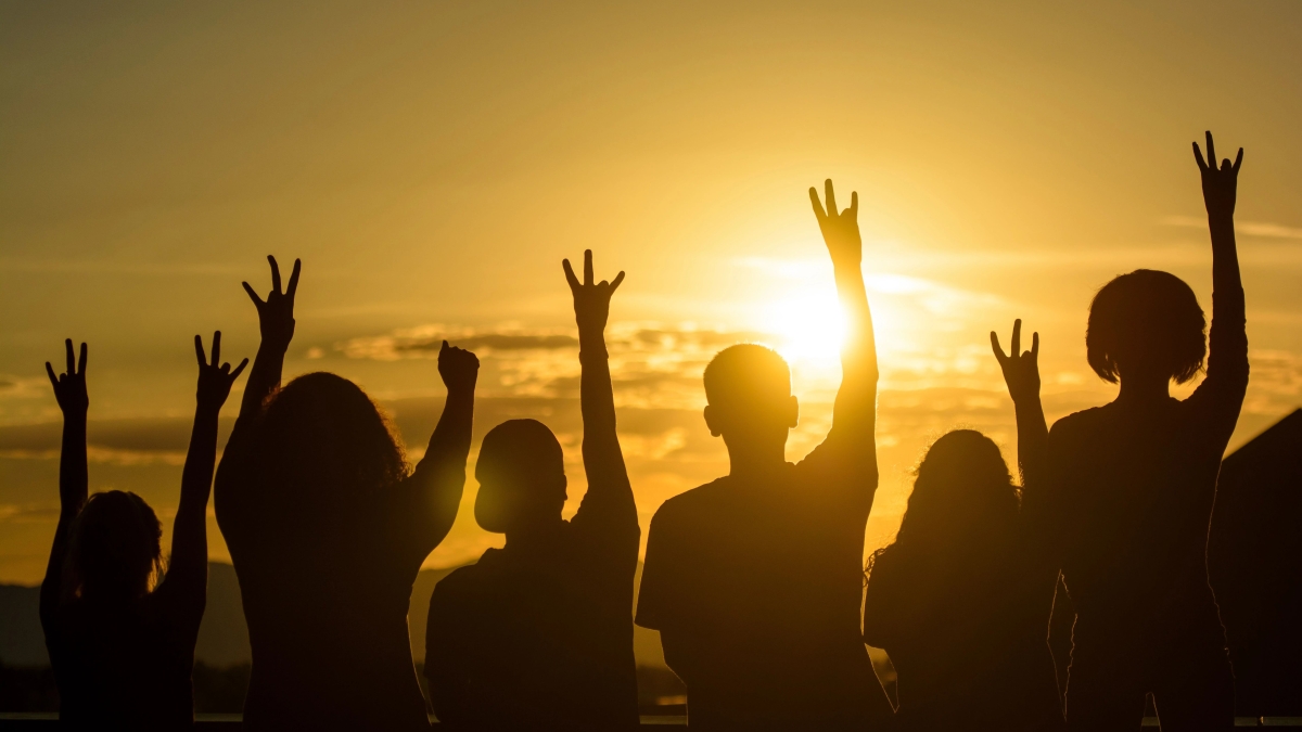 A group of people pictured from the back, silhouetted by a setting sun. They are raising their arms and making the pitchfork gesture with their hands.