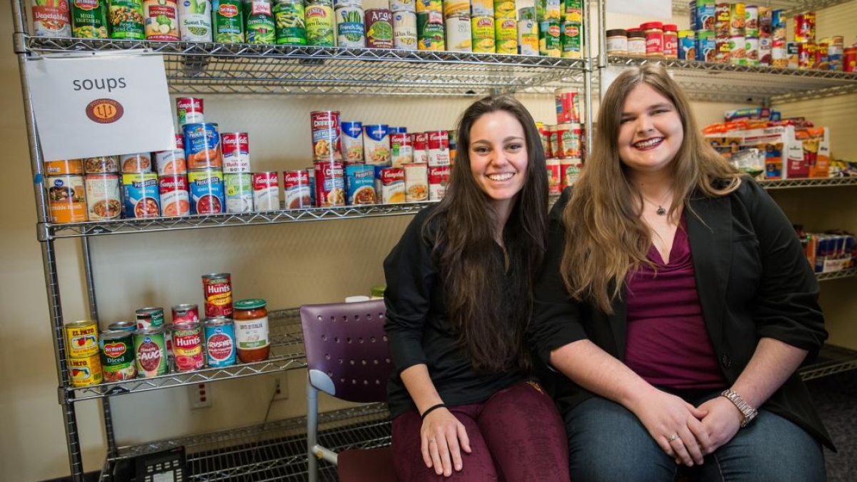 two women seated in front of shelves of canned food