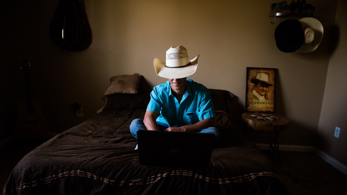 A boy in a cowboy hat sits on a bed and types on a laptop