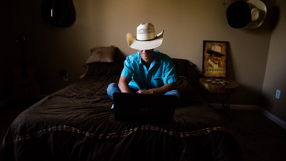 A boy in a cowboy hat sits on a bed and types on a laptop