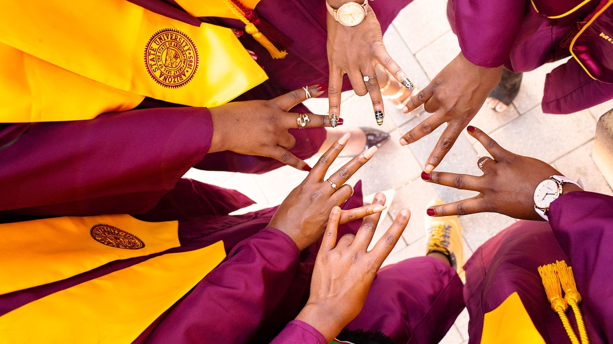A bunch of hands showing the ASU pitchfork gesture in the center of a circle of people wearing graduation gowns whose faces aren't seen 