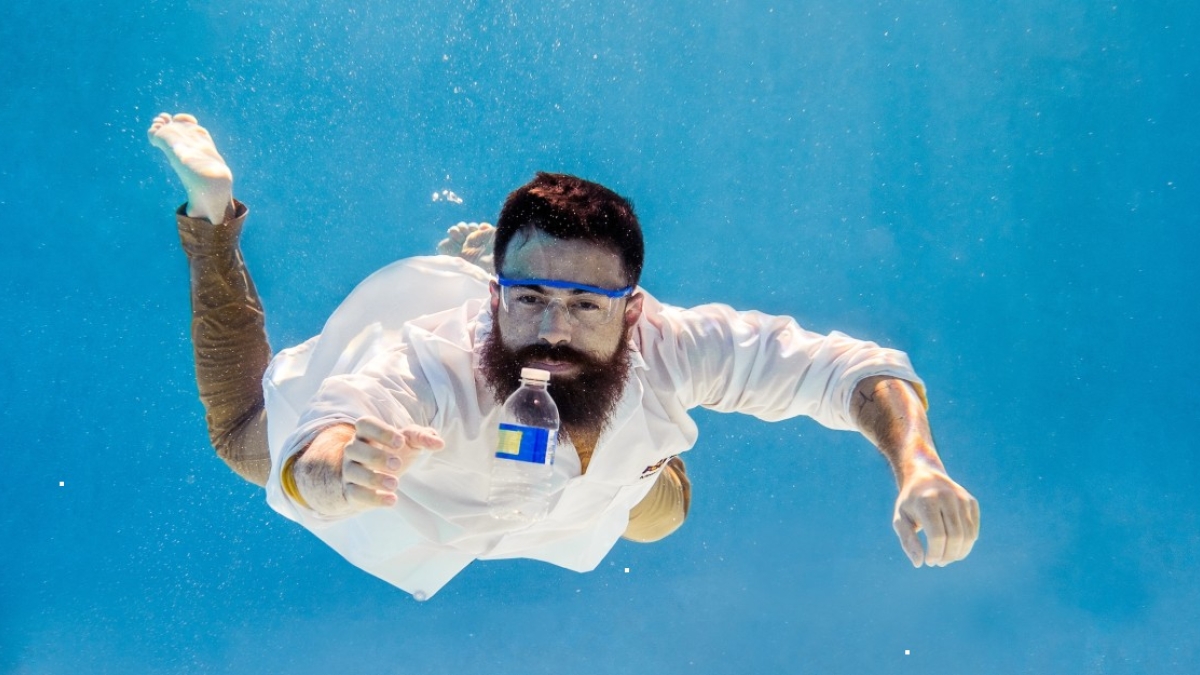 A man in a lab coat is suspended underwater, reaching out for a plastic water bottle in front of him.