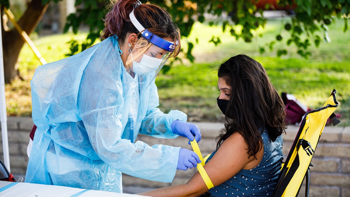 A woman in full personal protective gear prepares for a blood draw on another woman's arm.