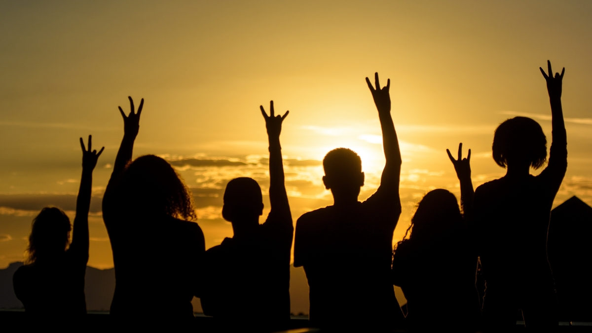 Silhouettes of a group of people in front of a sunset holding their hands in the air in the shape of an ASU pitchfork.