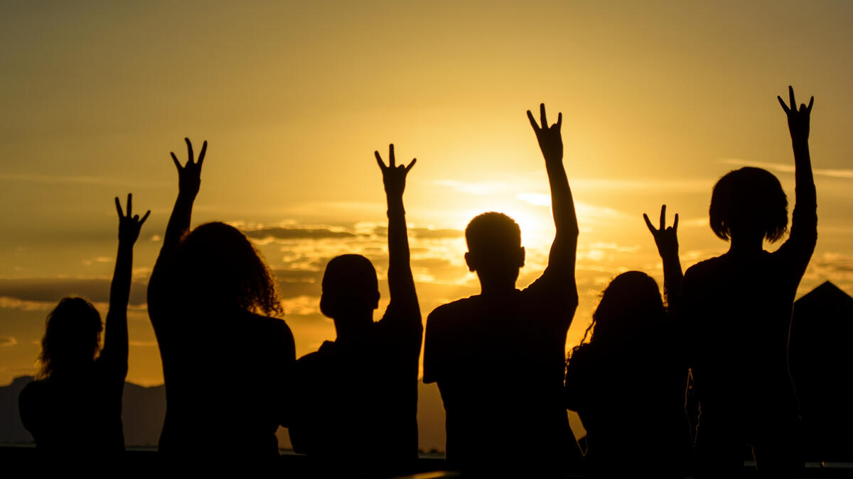 Group of people silhouetted against a sunset, each making the ASU pitchfork symbol with their hands.