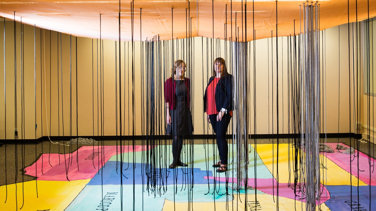 two women standing on exhibit of AZ map on floor with black strings hanging from ceiling