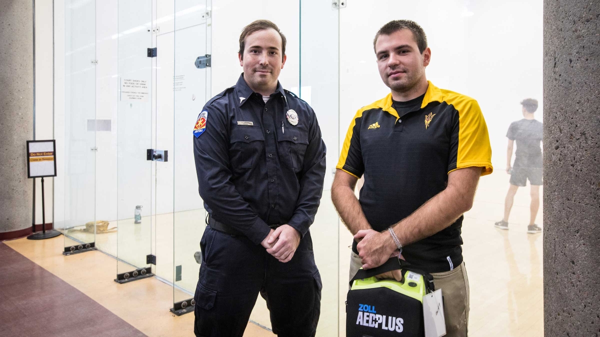 Two men stand in front of a racquetball court with CPR equipment