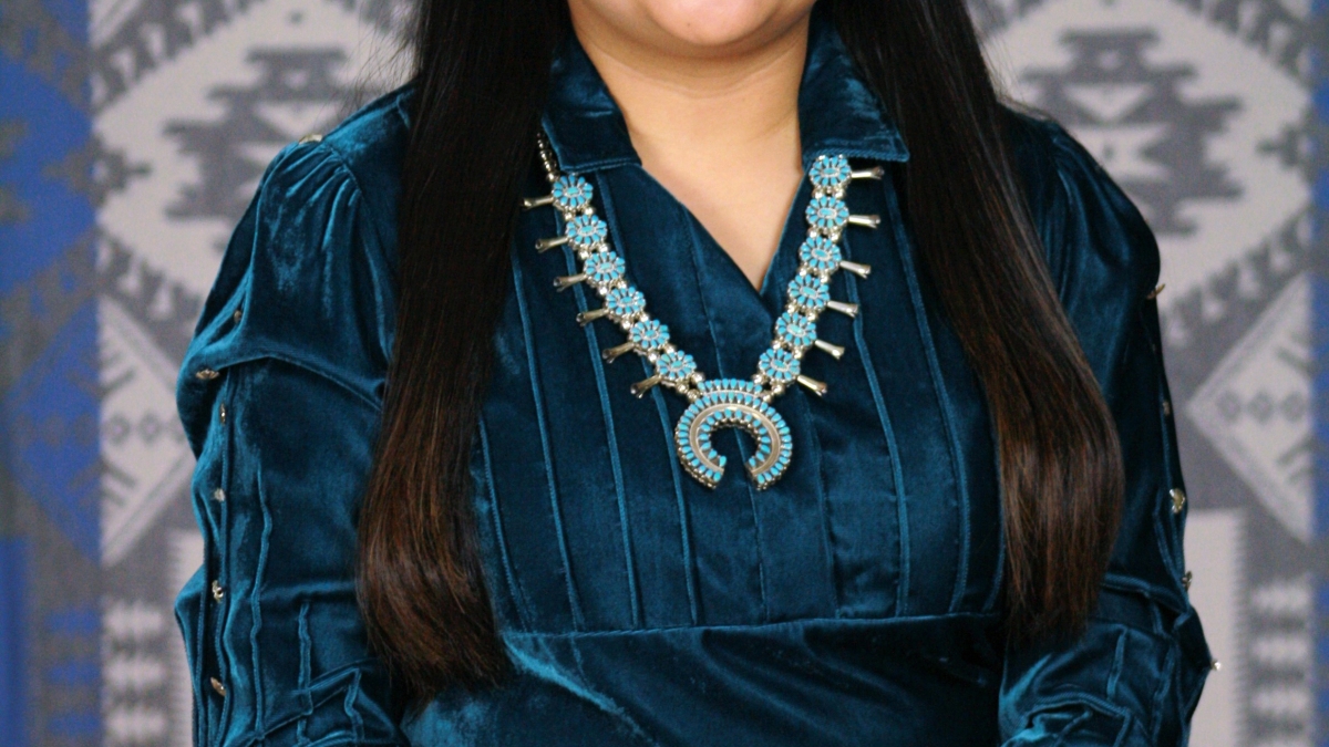 Jennifer Harrison (medium complexion with long dark hair, wearing native american turquoise jewelry and a dark teal dress) sits before a gray and white woven Native American tapestry