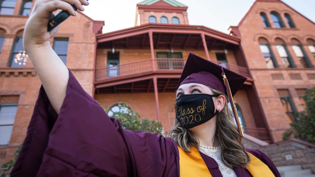 A student in graduation garb and a Class of 2020 mask takes a selfie in front of Old Main