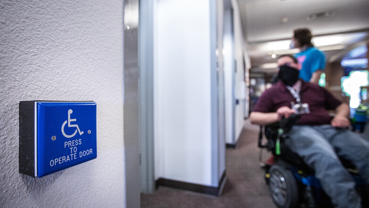 Wheelchair button access for bathroom in foreground, with man in wheelchair by door in background