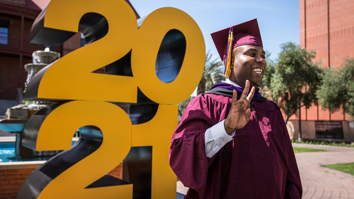 A man in a graduation cap and gown flashes the pitchfork gesture and smiles for a photo in front of a giant 2021 sign