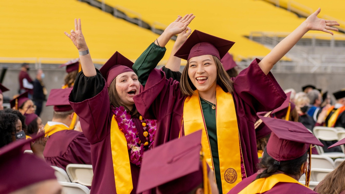 Two women in graduation gowns throw their arms up and cheer at graduation