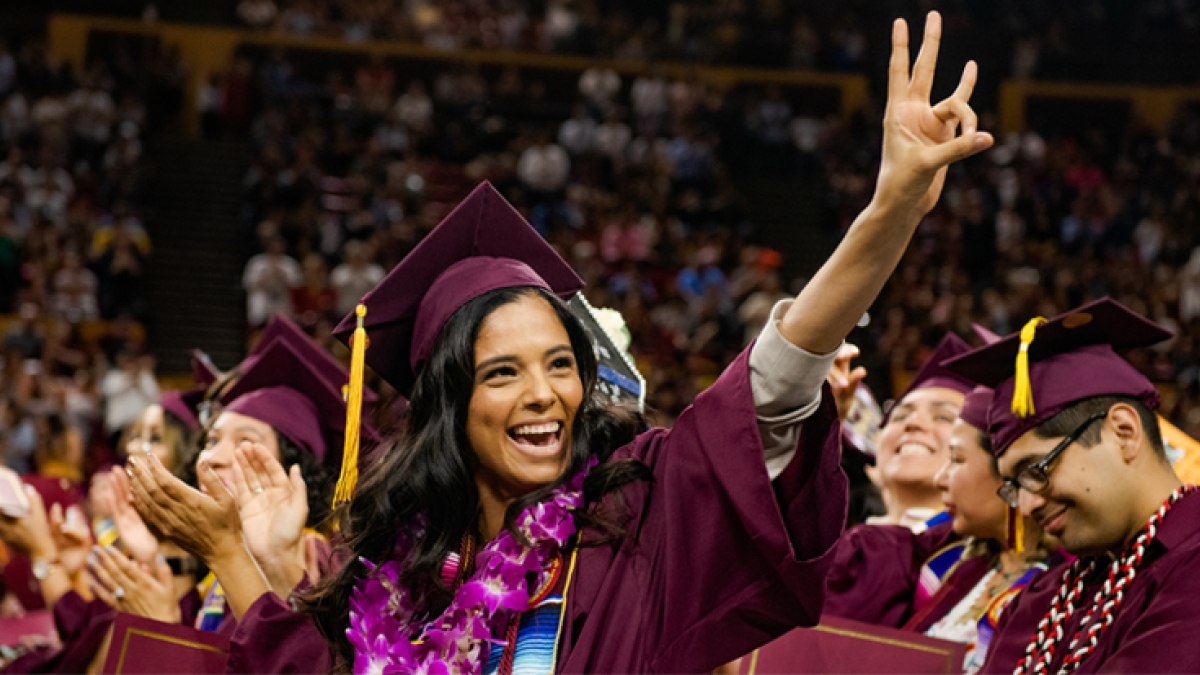Woman wearing cap and gown at an ASU graduation ceremony. She is smiling widely and making the pitchfork gesture.