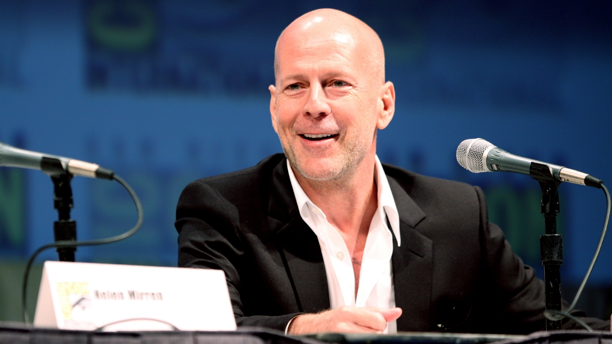 Actor Bruce Willis speaking into a microphone while seated at a table.