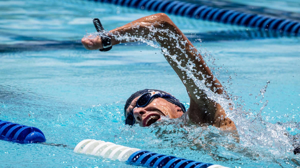 Man wearing a swim camp and goggles takes a breath mid-stroke as he swims down a lane in a pool.