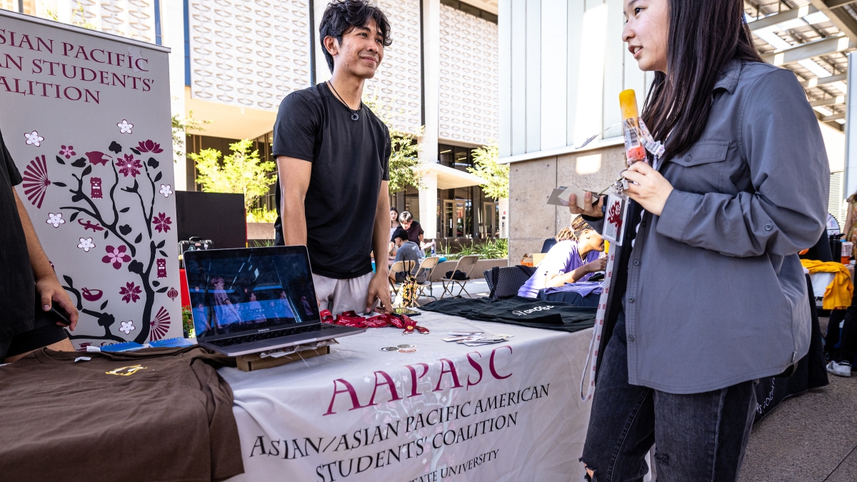 Student standing behind a table at the visiting booth speaking to another student with a popsicle at ASU's Culture Kickoff event.
