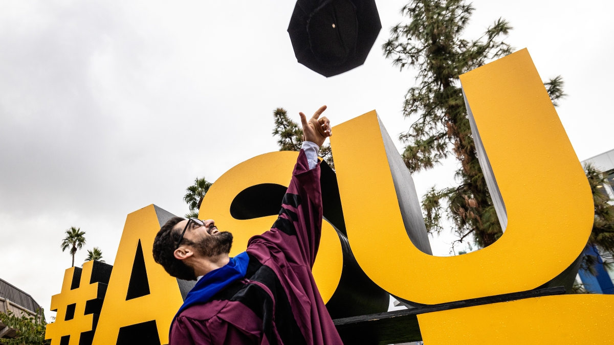 Graduate tossing a cap into the air in front of a gold "ASU GRAD" sign.