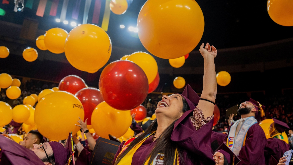 A woman in a graduation cap and gown smiles toward the ceiling as giant balloons drop down onto the graduates