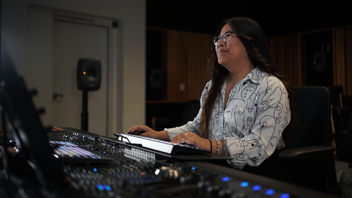 A smiling female student sits at a sound mixing board.