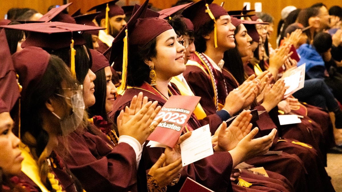 Students in caps and gowns clap while seated in an auditorium.