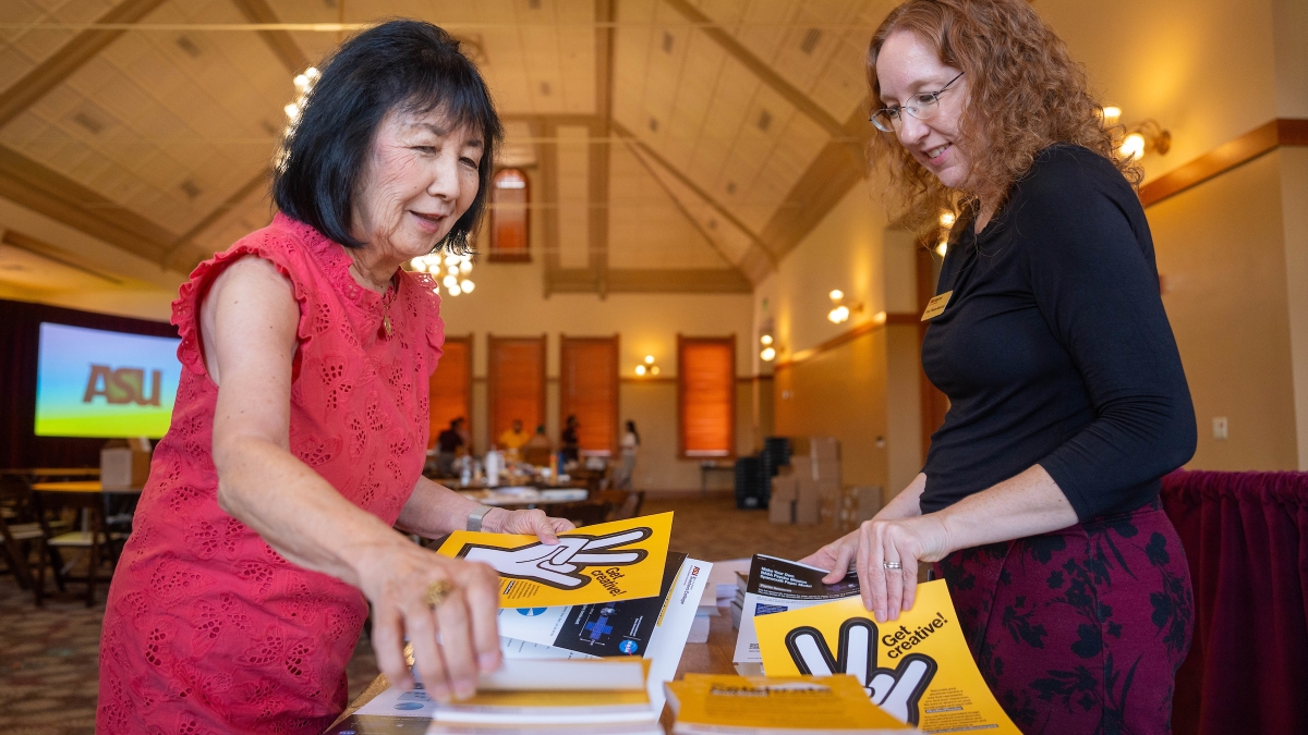 Two women reaching for flyers on a table.